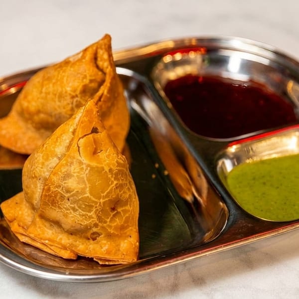 Two golden-brown samosas and two small bowls of dipping sauces on a silver platter.