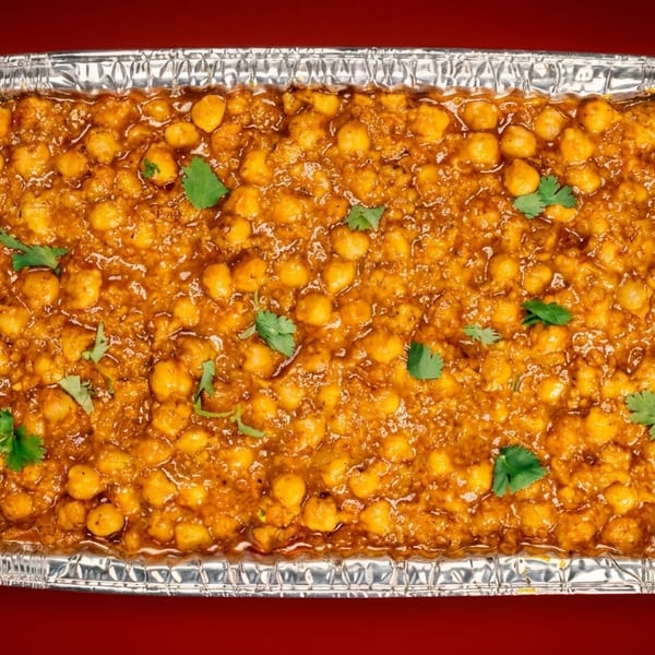 A rectangular foil tray filled with chana masala, garnished with fresh cilantro, on a red background.