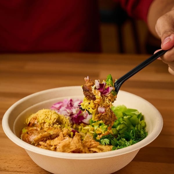 A hand holds a forkful of colorful Indian food from a bowl on a wooden table.