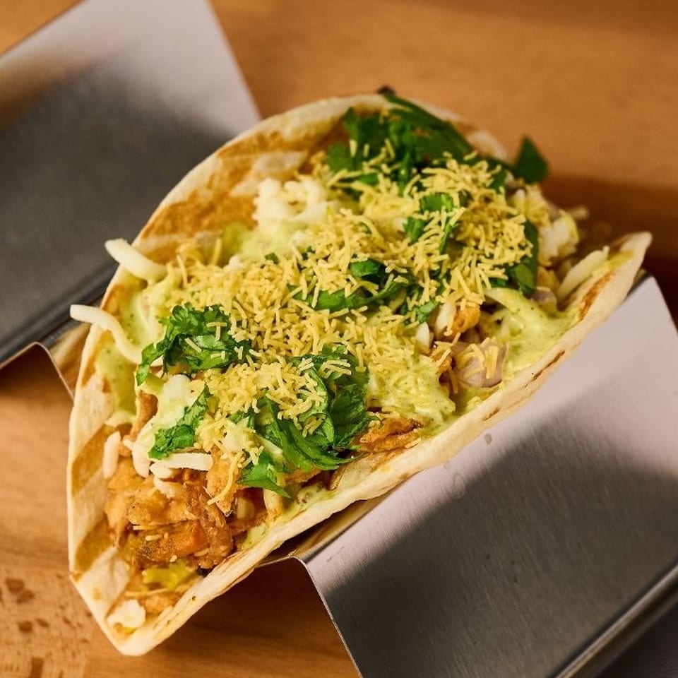 A taco filled with chicken, green sauce, cilantro, and crispy sev, presented in a metal holder.
