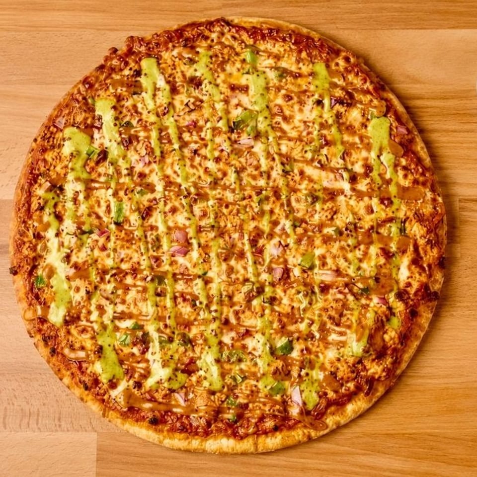 Whole pizza with green and dark sauce drizzle, cheese, onions, and peppers, beside a dipping cup.