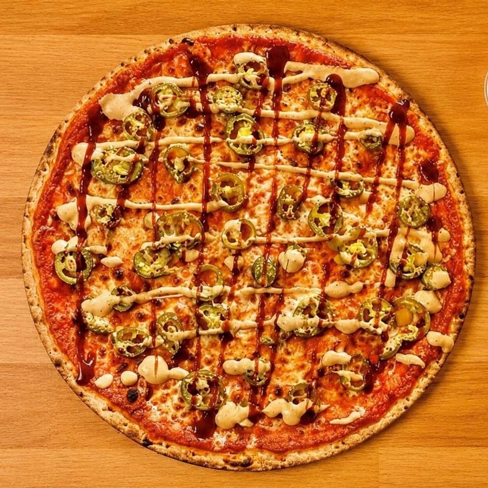 A whole pizza topped with jalapeños, cheese, BBQ sauce, and a creamy drizzle, with a side of dipping sauce.